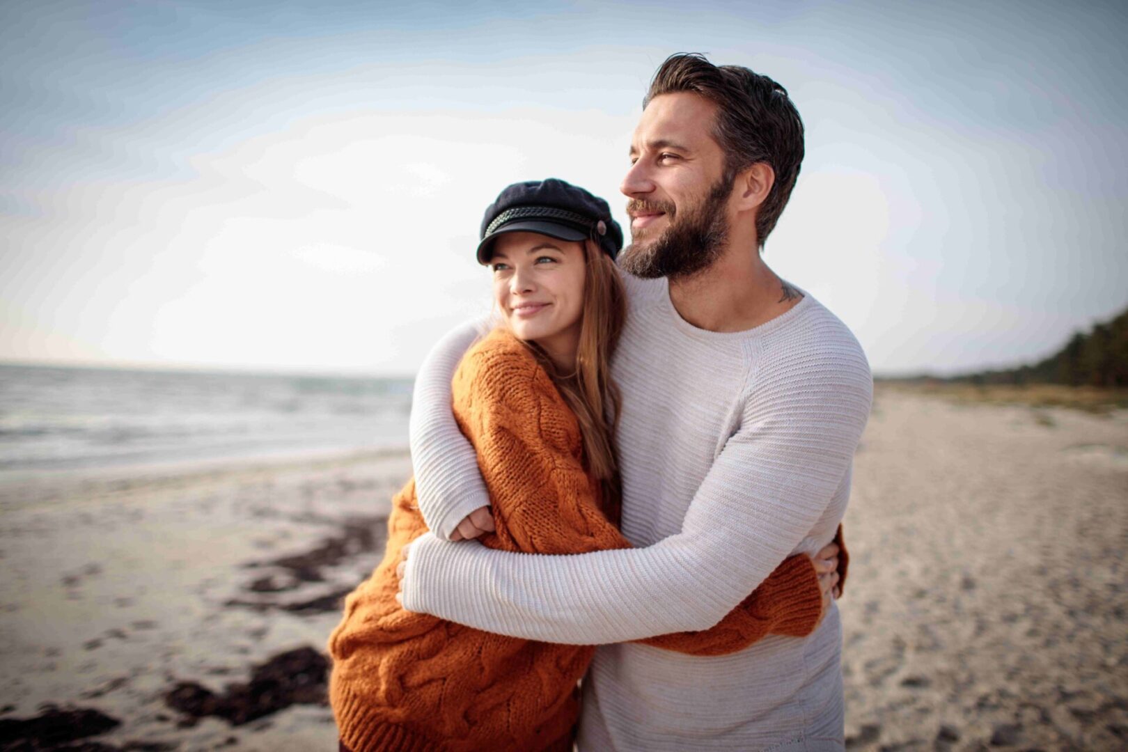 GettyImages-1081762006 A couple embraces on a beach, smiling. The woman wears an orange sweater and black cap, and the man wears a gray sweater. The sea and sky are visible in the background.
