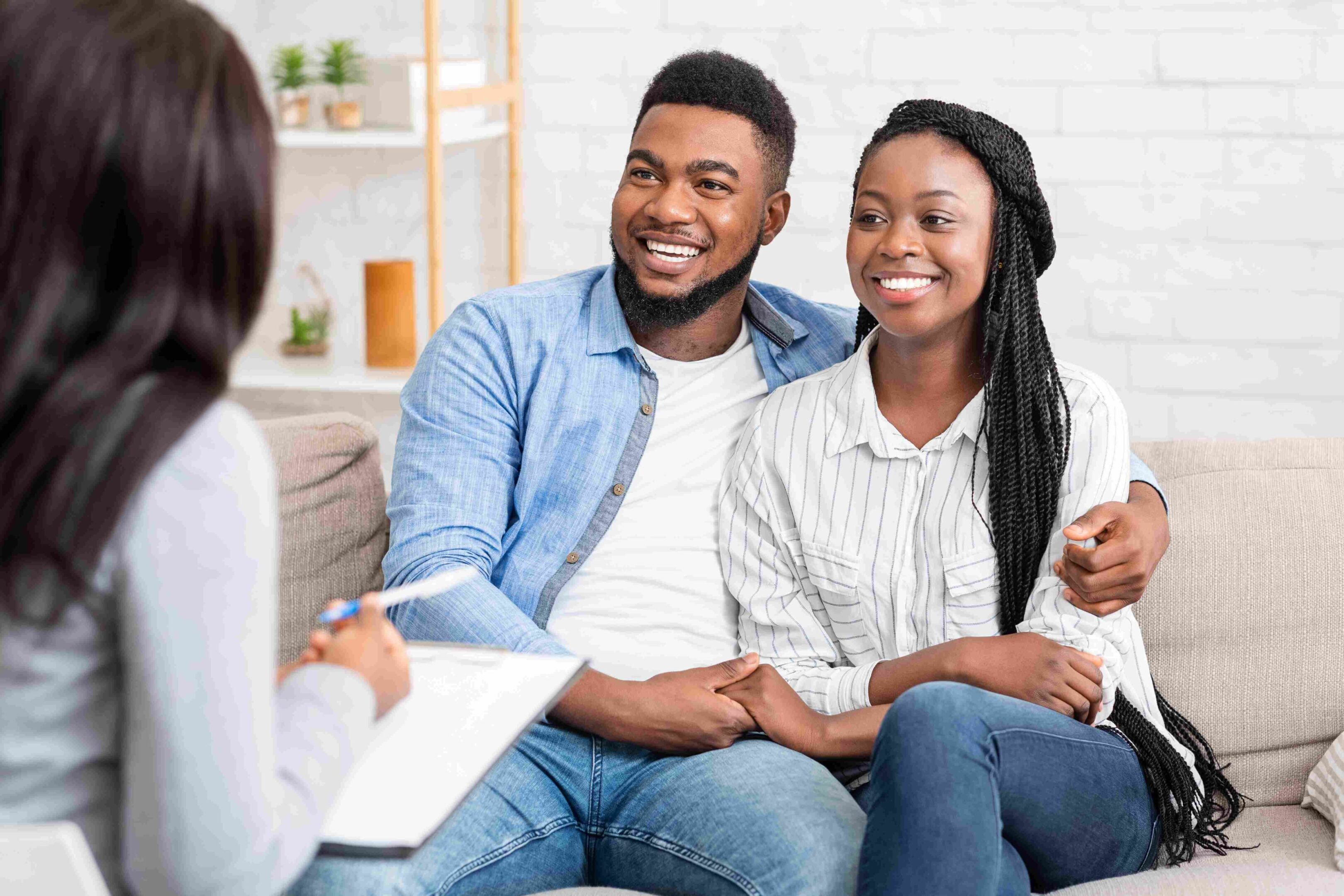 GettyImages-1191647689 (1) (1) A couple sits on a sofa, smiling and holding hands during their couples therapy session. Engaging warmly with the counselor, who holds a clipboard and pen, they seem hopeful and connected as they navigate this journey together.