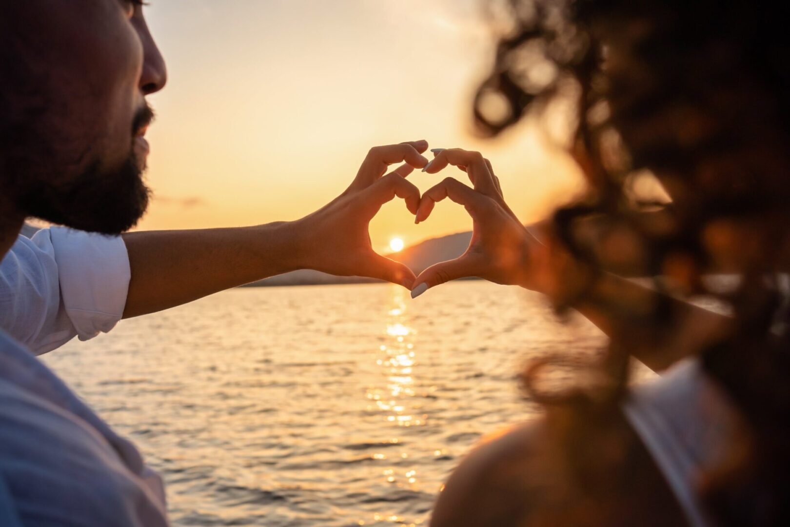 GettyImages-1299412800 Against the backdrop of a stunning sunset over the water, two people form a heart shape with their hands, symbolizing unity and commitment—a perfect reflection of The Brave Marriage mission.