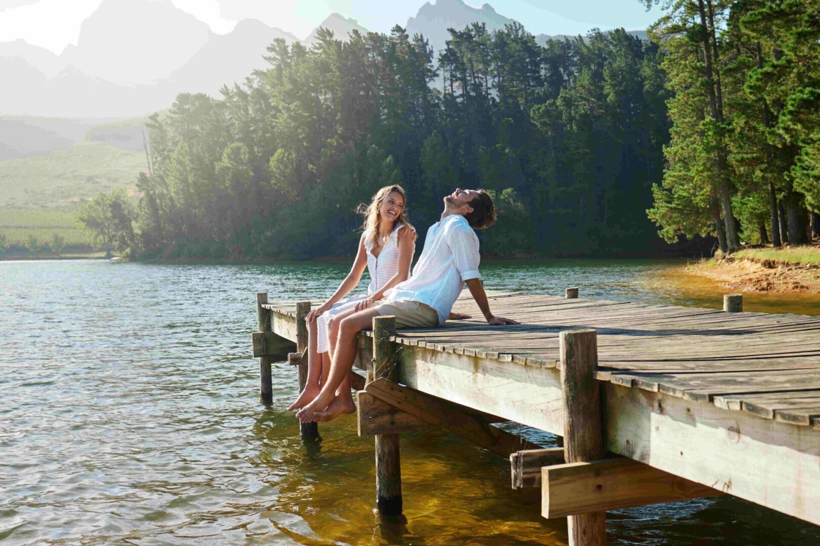 GettyImages-1391730246 A man and woman sit on a wooden dock, smiling by a calm lake surrounded by trees and mountains on a sunny day, perhaps reflecting on insights from couples counseling that brought them closer together.