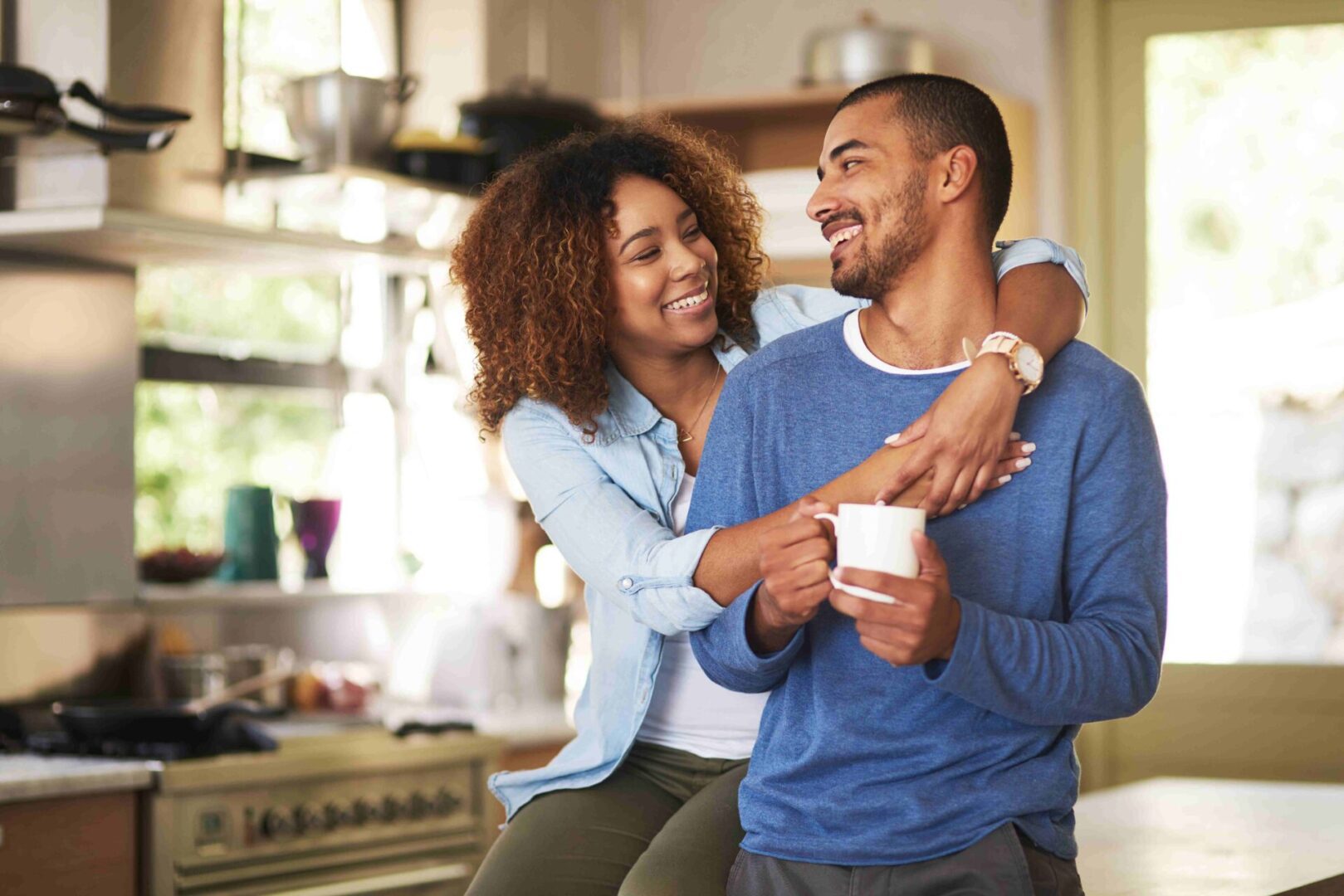 GettyImages-639583980 A woman embraces a smiling man sitting on a kitchen counter, holding a coffee mug. Both appear cheerful in a well-lit kitchen setting.