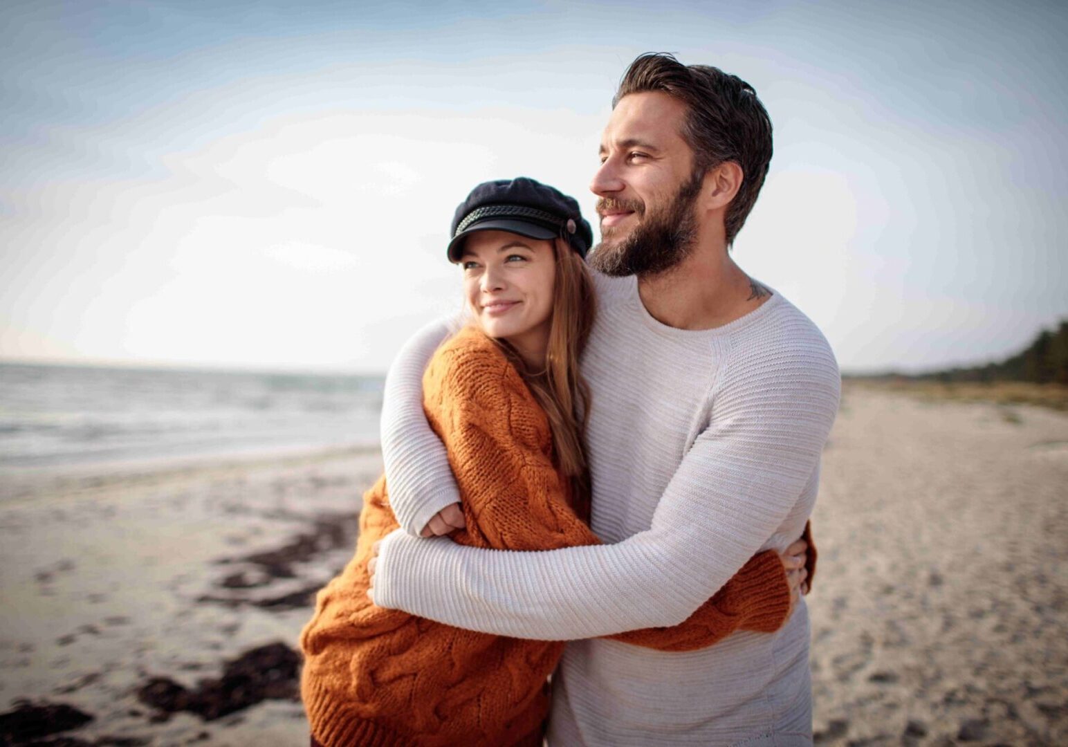 A couple embraces on a beach, smiling. The woman wears an orange sweater and black cap, and the man wears a gray sweater. The sea and sky are visible in the background.