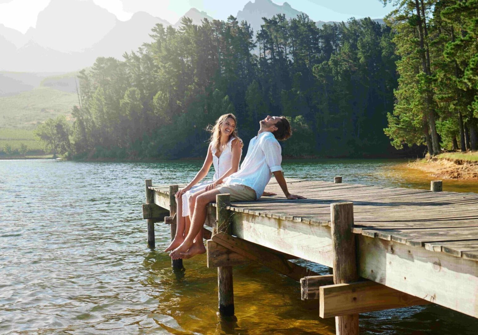 A man and woman sit on a wooden dock, smiling by a calm lake surrounded by trees and mountains on a sunny day, perhaps reflecting on insights from couples counseling that brought them closer together.