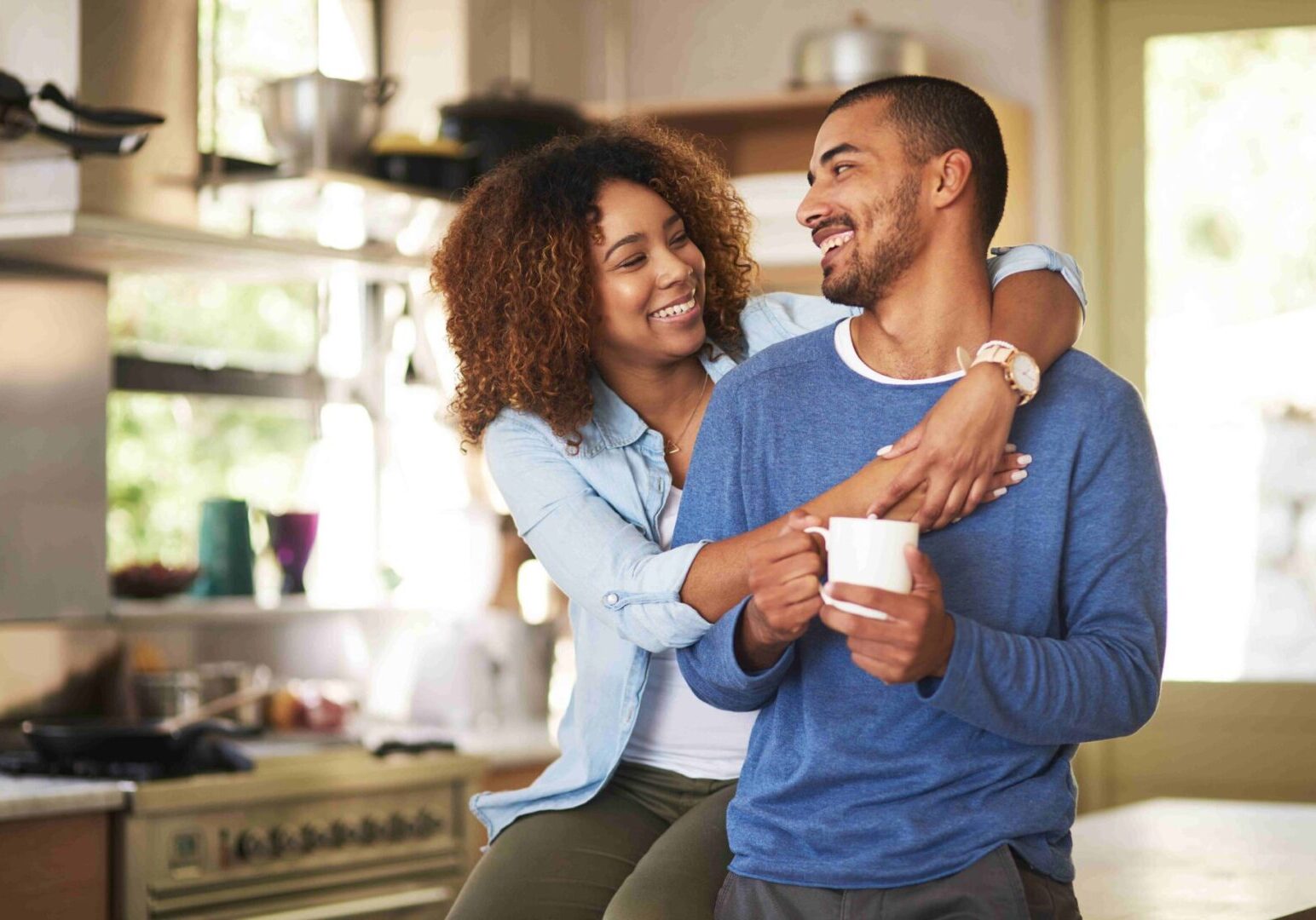 A woman embraces a smiling man sitting on a kitchen counter, holding a coffee mug. Both appear cheerful in a well-lit kitchen setting.
